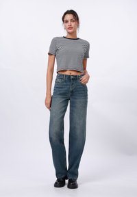 Ženska v črno-beli prugasti skrajšani majici, visokih modrih kavbojkah in črnih čevljih, stoji na enobarvni beli podlagi.