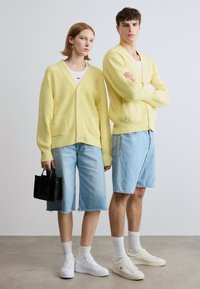Ανοιχτό κίτρινο πλεκτό ζακέτα φορεμένο πάνω από λευκές μπλούζες, συνδυασμένο με τζιν σορτς. Η μία κρατάει μια μικρή μαύρη τσάντα χειρός· και οι δύο φορούν λευκά αθλητικά παπούτσια και κάλτσες.