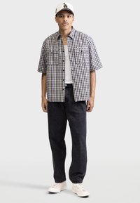 Schwarz-weiße karierte Kurzarmbluse mit zwei vorderen Taschen, kombiniert mit dunklen Jeanshosen und weißen Sneakers.