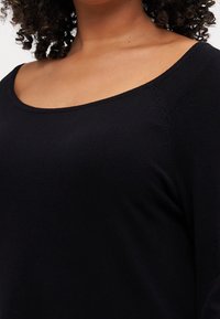 Vrouw met krullend haar in een zwarte top met een ronde hals, die de schouders en het bovenste deel van de borst toont.