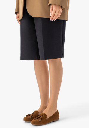 Osoba ubrana w czarne spodenki do połowy uda, brązową marynarkę oraz brązowe zamszowe mokasyny z chwostami na białym tle.