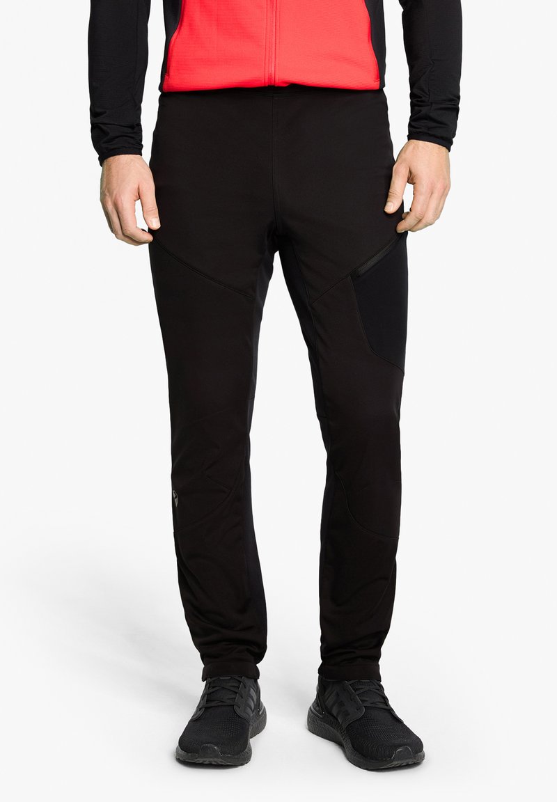 Schwarze Sporthose mit einer figurschmeichelnden Passform, ausgestattet mit Kniepaneelen, Reißverschlusstaschen und einer glatten Textur. Kombiniert mit schwarzen Schuhen.