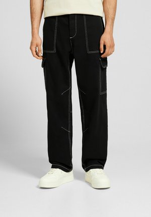 Mand iført sorte cargobukser med hvide syninger og hvide sneakers, stående mod en ensfarvet lys baggrund.
