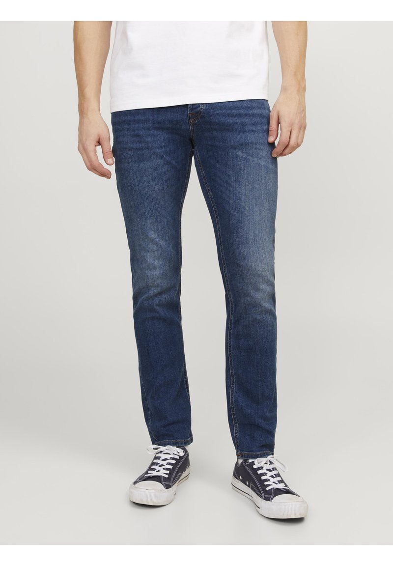Mann, der schlanke, blaue Jeans und marineblaue Canvas-Sneaker trägt, steht vor einem schlichten, hellen Hintergrund.