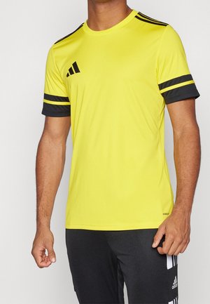 Homme portant un t-shirt de sport Adidas jaune vif avec des rayures noires sur les manches et un pantalon Adidas noir avec des rayures blanches.
