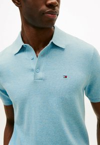 Polo de color azul claro hecho de tejido texturizado, con una tapeta de dos botones y un pequeño logo bordado en rojo y azul marino en el pecho.