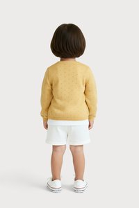 Ein Kind steht mit dem Rücken zur Kamera und trägt einen senfgelben Pullover mit einem kleinen Lochmuster, weiße Shorts und weiße Turnschuhe vor einem einfarbigen Hintergrund.