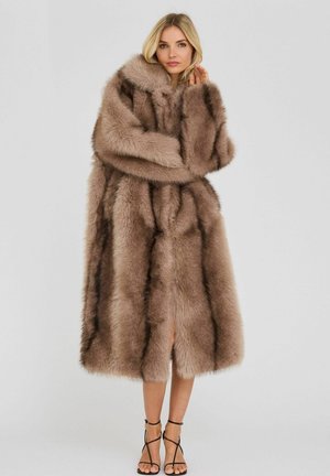 Femme blonde porte un long manteau épais en fourrure beige avec des rayures foncées, debout les bras croisés, assorti à des talons hauts noirs à brides.
