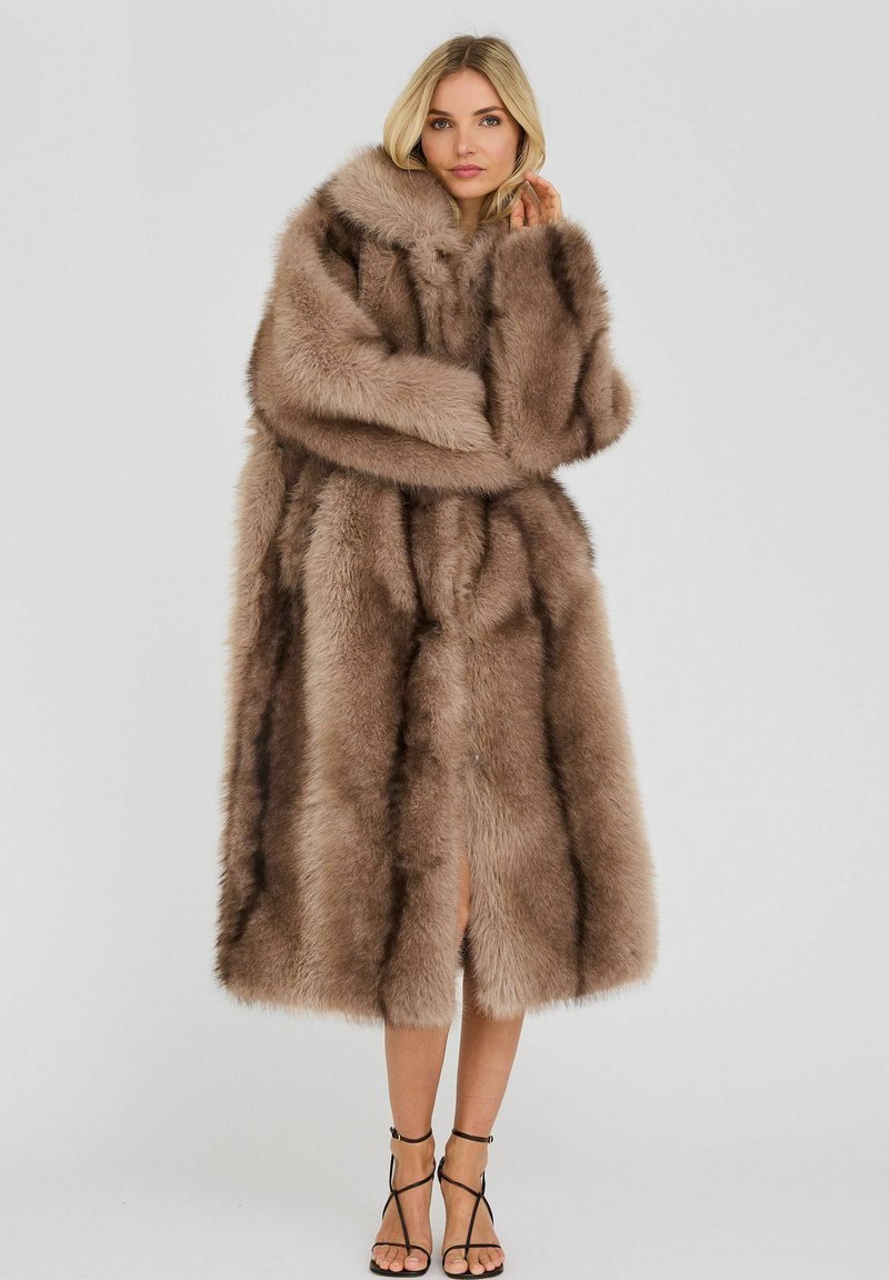 Femme blonde porte un long manteau épais en fourrure beige avec des rayures foncées, debout les bras croisés, assorti à des talons hauts noirs à brides.