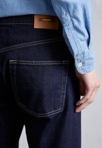 Dark blue denim jeans with yellow stitching, showing a back pocket. A light blue shirt cuff is partially visible, with a button detail.