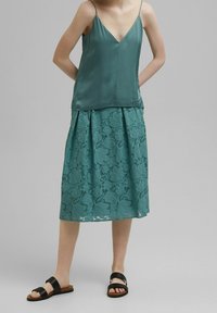 Camiseta de satén verde azulado con tirantes finos, combinada con una falda de encaje verde azulado con patrones florales. Sandalias planas negras completan el conjunto.
