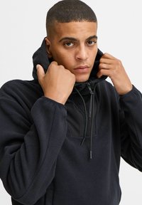 Svart fleece genser med halv glidelås foran og hette i kontrasterende svart stoff. Har en teksturert overflate og justerbar snor.