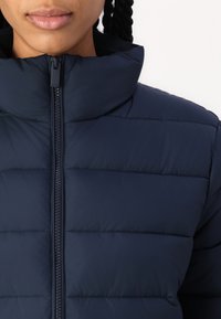 Geacă puffer de culoare bleumarin, cu guler înalt, panouri quilted orizontale și fermoar frontal pentru închidere. Textură moale, material ușor.