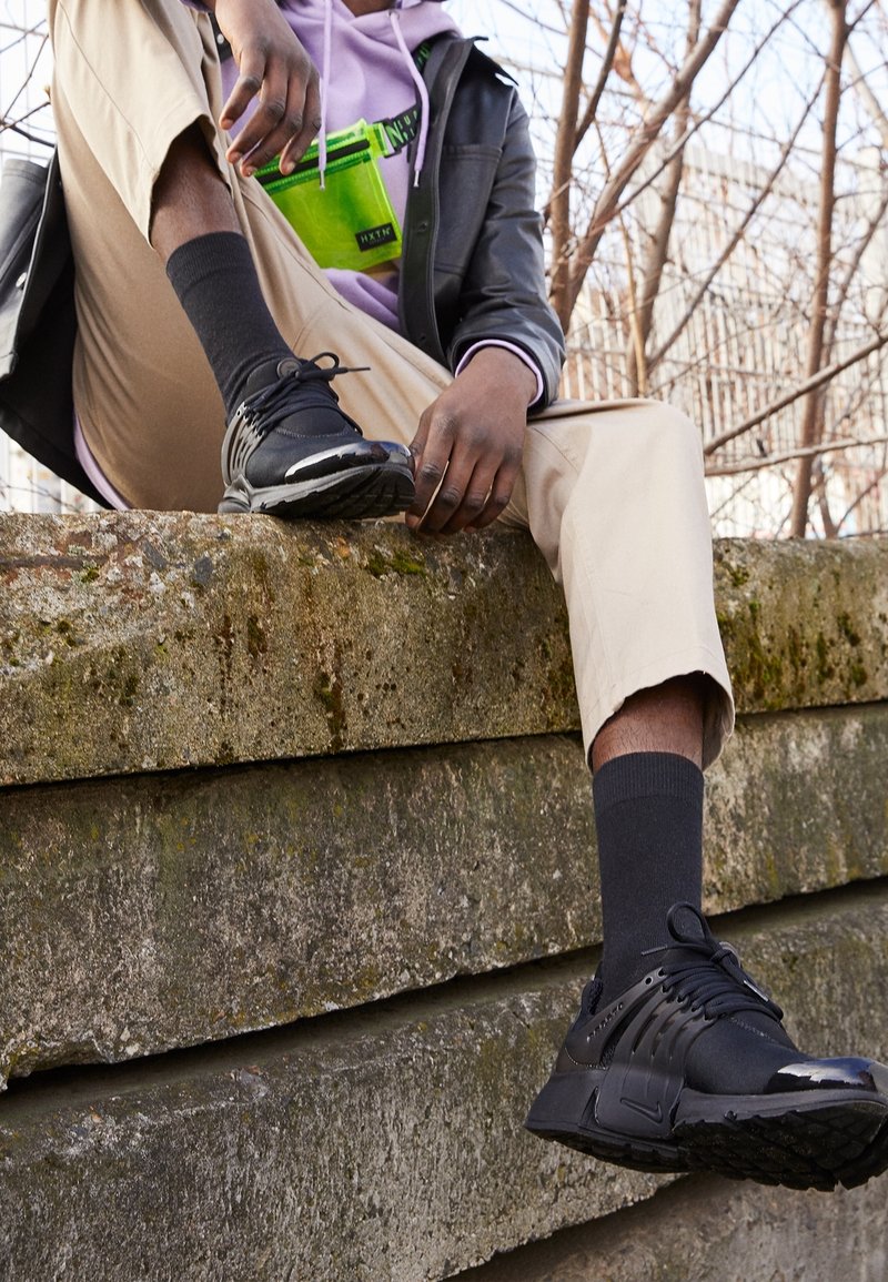 Osoba v béžových kalhotách, černých teniskách, černých ponožkách, fialové mikině a černé bundě sedí na opotřebované betonové římse venku.