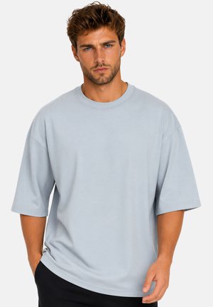 Uomo con capelli corti castani e barba che indossa una camicia oversize grigio chiaro a maniche corte e pantaloni neri su uno sfondo semplice.