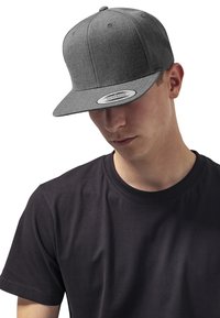 Casquette snapback grise avec une visière plate, en tissu texturé, dotée de six panneaux et d'une étiquette intérieure de marque. Portée bas sur un t-shirt noir.