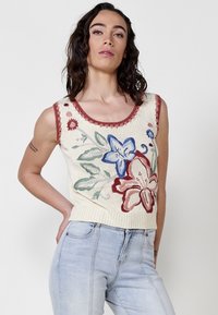 Suéter sin mangas de punto en color crema con bordados florales en rojo, azul y verde. Presenta un escote con forma de ondas y un dobladillo acanalado.