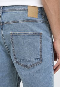 Hele sinised teksapüksid, millel on üks tagatasku kontrastsete õmblustega ja vöökohal beež nahast märk.