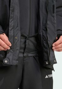 Schwarze Jacke mit einem strukturierten Innenfutter und Druckknopfverschlüssen, ausgestattet mit einem Reißverschluss vorne und elastischen Bündchen. Kombiniert mit schwarzen Hosen.