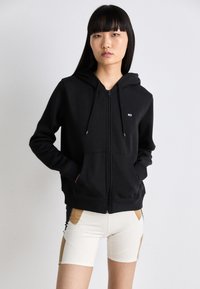 Hoodie zippé noir avec cordons, présentant un patch logo sur la poitrine gauche et deux poches avant. Tissu lisse et coupe relaxe.