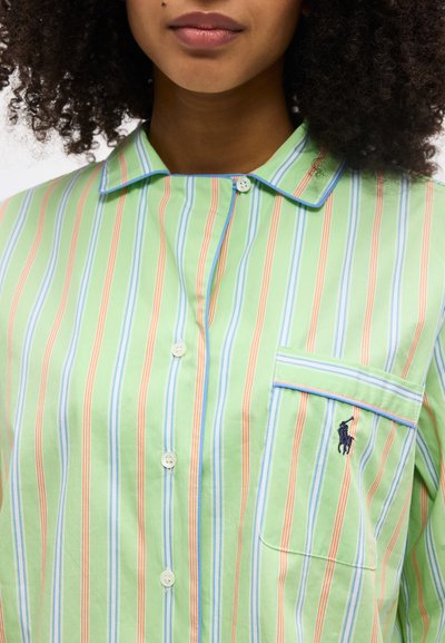 Γυναίκα που φοράει ένα ανοιχτοπράσινο πουκάμισο με πορτοκαλί, μπλε και λευκές κάθετες ρίγες και μια τσέπη που φέρει ένα μικρό κεντημένο λογότυπο.