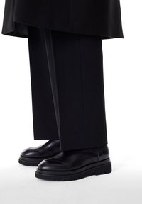 Zapatos de cuero negros con punta redondeada, con una superficie texturizada y suela de goma gruesa. Combinados con pantalones negros.