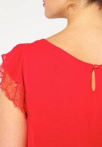 Blouse rouge avec bordure en dentelle sur les manches courtes à volant et une ouverture en trou de serrure à l'arrière, confectionnée dans un tissu lisse et léger.