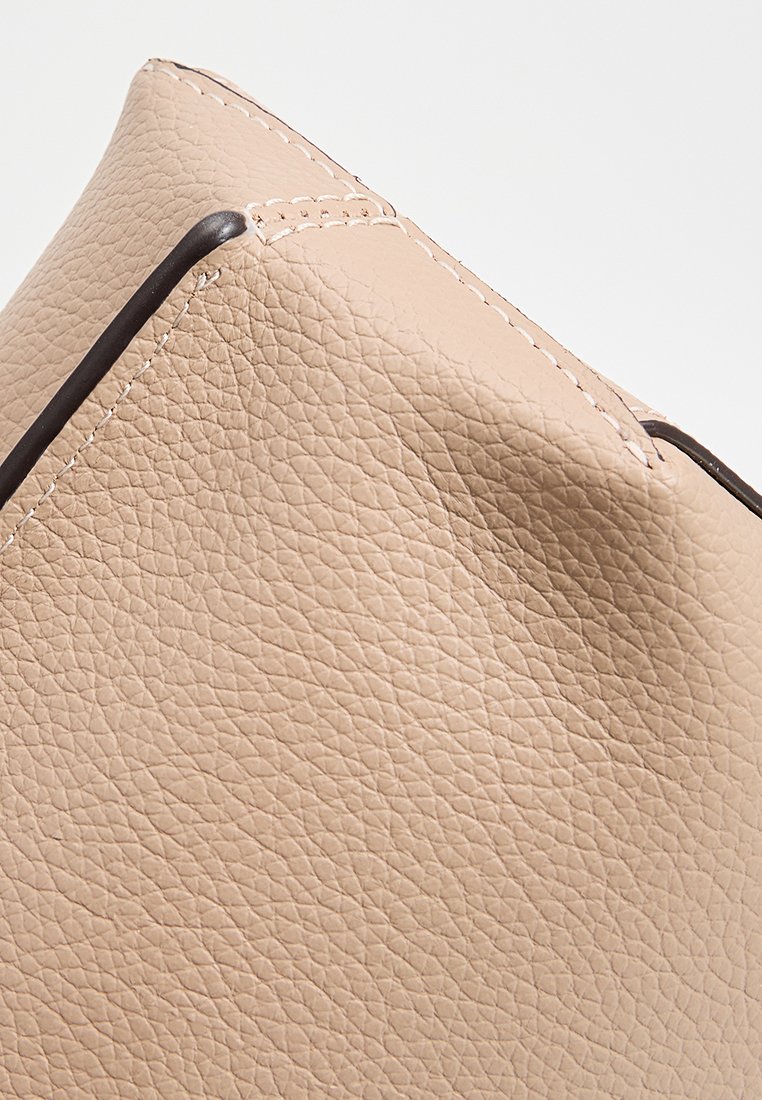 Coin de sac à main en cuir beige avec une surface texturée, une bordure foncée contrastante et des détails de couture visibles. Forme angulaire.