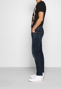 Tmavě modré džínové kalhoty slim fit s jemným vyblednutím, v kombinaci s černým tričkem s barevným květinovým grafikou a bílými teniskami.