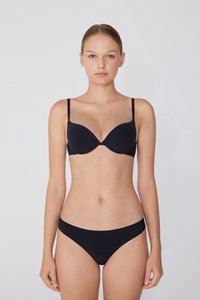 Mladá žena se světlou pletí, s vlasy staženými dozadu, oblečená v černé podprsence a odpovídajících černých bikinách, stojící na jednobarevném bílém pozadí.