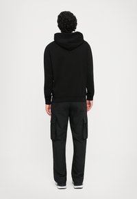 Homem de costas, vestindo um hoodie preto, calças cargo pretas e ténis preto e branco, contra um fundo claro e simples.