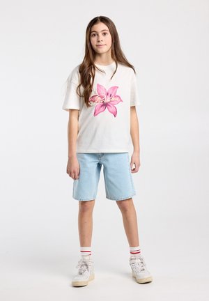 Jong meisje met lang bruin haar, gekleed in een wit t-shirt met een roze bloemmotief, spijkershorts, witte sneakers en gestreepte sokken.