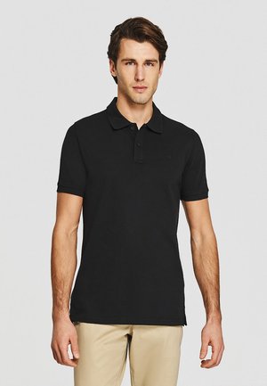 Mężczyzna z krótkimi brązowymi włosami, ubrany w czarną koszulkę polo z krótkim rękawem i beżowe spodnie, stojący na tle jednolitego jasnoszarego tła.