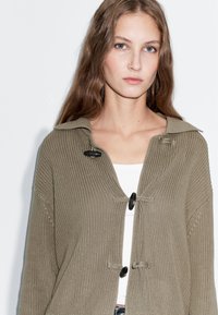 Olijfgroene gebreide cardigan met grote kraag, zwarte knoopsluitingen en golvende ribbeldetails. Draag over een wit shirt.