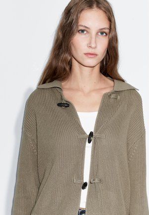 Cardigan verde oliva lavorato a maglia con grande colletto, chiusure con bottoni neri e dettagli a coste strutturate. Indossato sopra una maglietta bianca.
