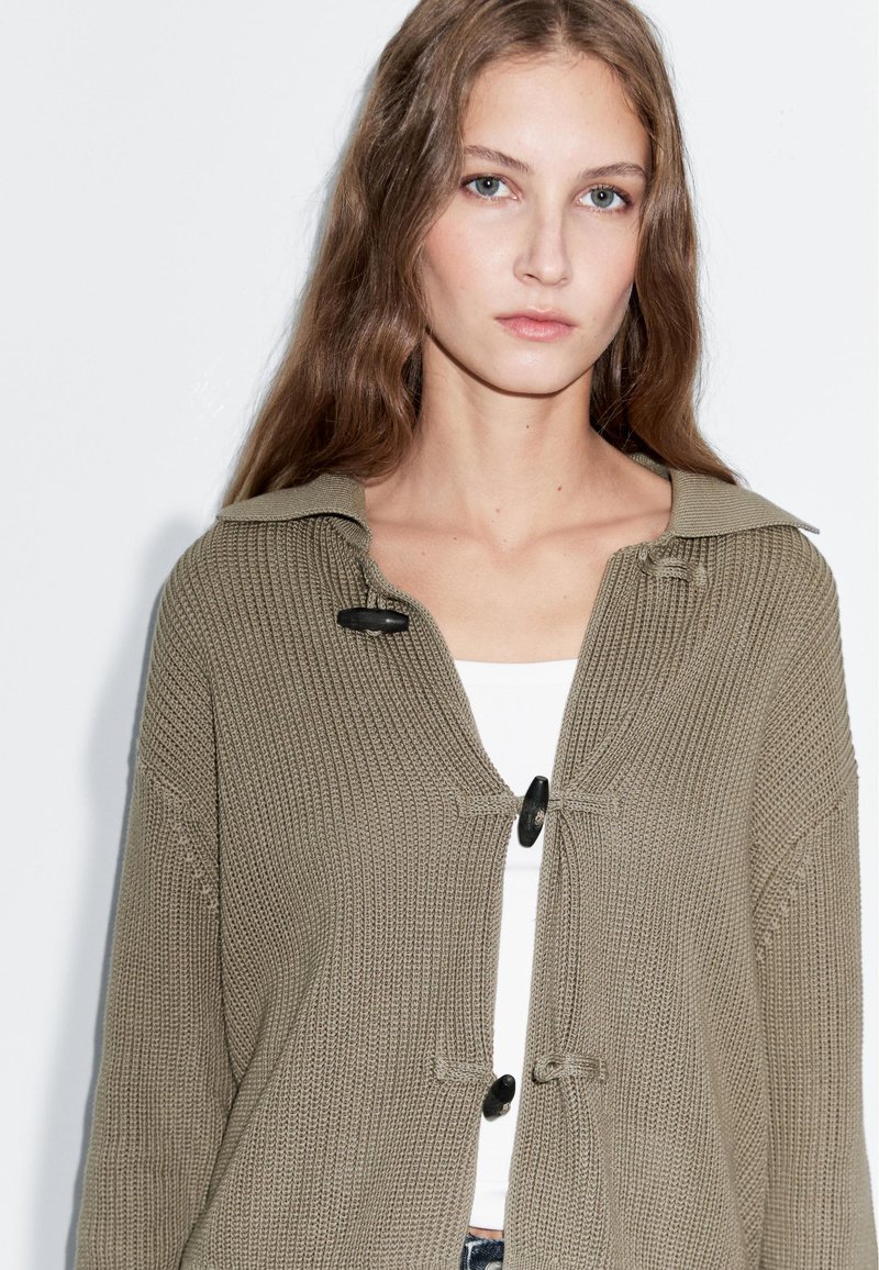 Oliwkowo-zielony dziergany kardigan z dużym kołnierzem, czarnymi guzikami i teksturowanymi prążkowaniami. Noszony na białej bluzce.