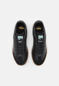 Sneaker in pelle e suede nera con suola in gomma, dotate di lacci neri e di un logo Puma verde e bianco sulla linguetta.