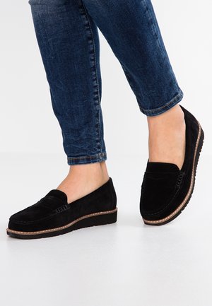 Schwarze Wildleder-Slipper mit einer gesteppten Akzentnaht und einer niedrigen Plateausohle, kombiniert mit einer dunklen Jeans.