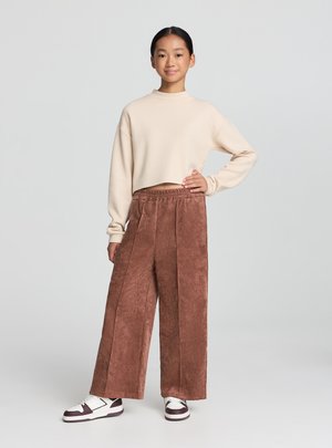 Felpa beige corta con maniche lunghe, abbinata a pantaloni marroni in velluto a coste a gamba larga. L'outfit è completato da sneakers bianche e marroni.