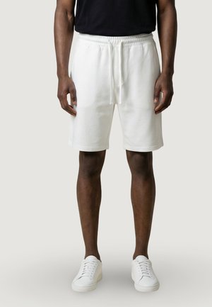 Mann trägt weiße Shorts mit Kordelzug, weiße Turnschuhe und ein schwarzes Hemd, steht vor einem schlichten hellen Hintergrund.