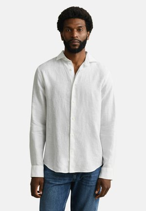 Homme portant une chemise blanche à manches longues boutonnée et un jean bleu, debout devant un fond clair uni.