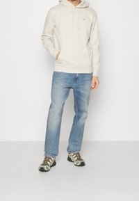 Persona che indossa un hoodie crema, jeans azzurri chiari e scarpe da trekking verdi, in piedi con una mano in tasca.