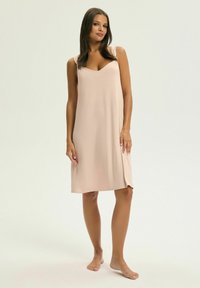 Bladoróżowa koszula nocna do kolan z dekoltem w serek i cienkimi ramiączkami, wykonana z miękkiego, gładkiego materiału. Brak widocznych wzorów lub detali metalowych.