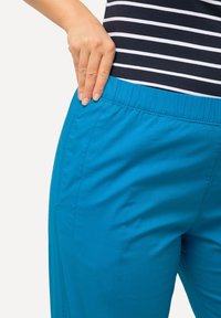 Een paar levendig blauwe broek met een elastische tailleband, voorzien van zijzakken en gemaakt van een gladde, lichte stof, weergegeven tegen een neutrale achtergrond.