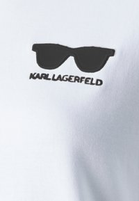 Balta medvilnės marškinėlių palaidinė su juodu saulės akinių grafiniu elementu ir užrašu "KARL LAGERFELD", abu kuriuose yra paprastas, tvarkingas dizainas.