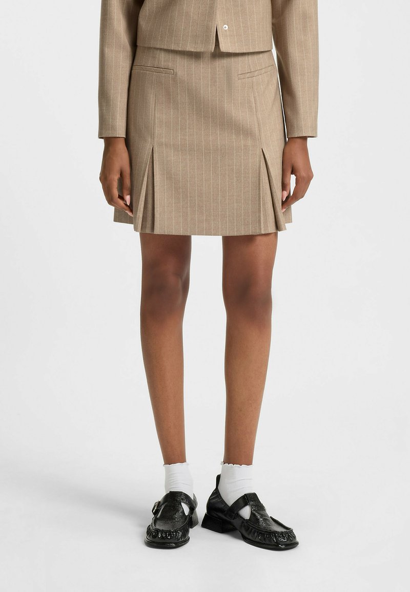 Beige hame pinstriped dengan lipit di bagian bawah, kantong depan, dan potongan yang disesuaikan. Dipadukan dengan sepatu hitam dan kaus kaki pergelangan putih.