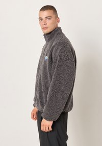 Uomo che indossa una giacca in pile grigia con collo alto e zip, con una superficie testurizzata e soffice, in piedi di lato contro uno sfondo chiaro e uniforme.