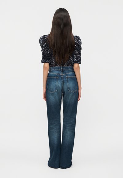 Γυναίκα με μακριά σκούρα μαλλιά, φοράει μαύρη μπλούζα με σχέδιο και ψηλόμεσα ξεθωριασμένα μπλε τζιν παντελόνια με ίσια μπατζάκια, στέκεται με την πλάτη προς τα εμάς.