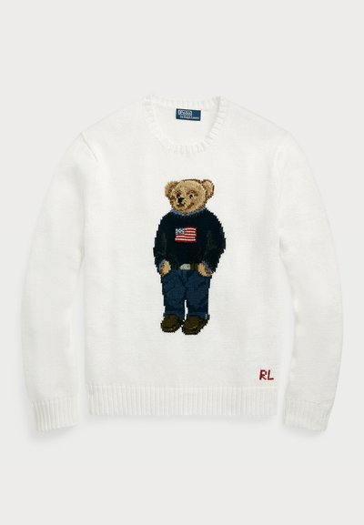 Sweater branco tricotado com um gráfico de um urso a usar um suéter azul-marinho com uma bandeira americana, jeans azuis e sapatos castanhos. "RL" em vermelho na bainha.