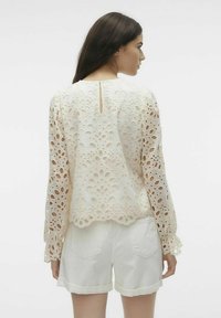 Blonde bluse med lange ermer, kremfarge, med intrikate utskjæringsmønstre og bølgekant. Sammen med hvite shorts.